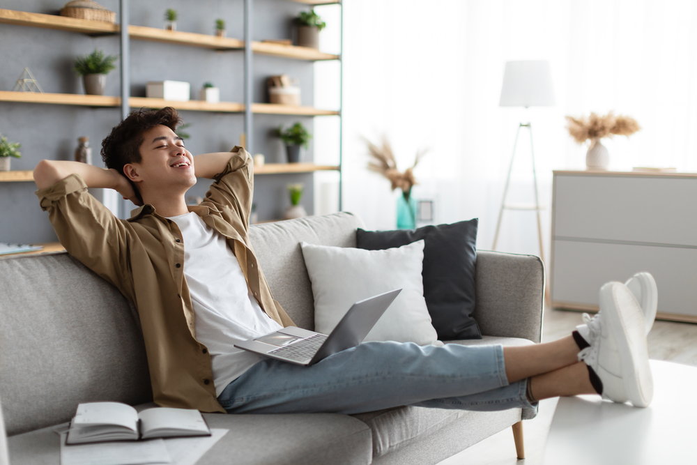 Smiling asian man resting on couch with pc on lap